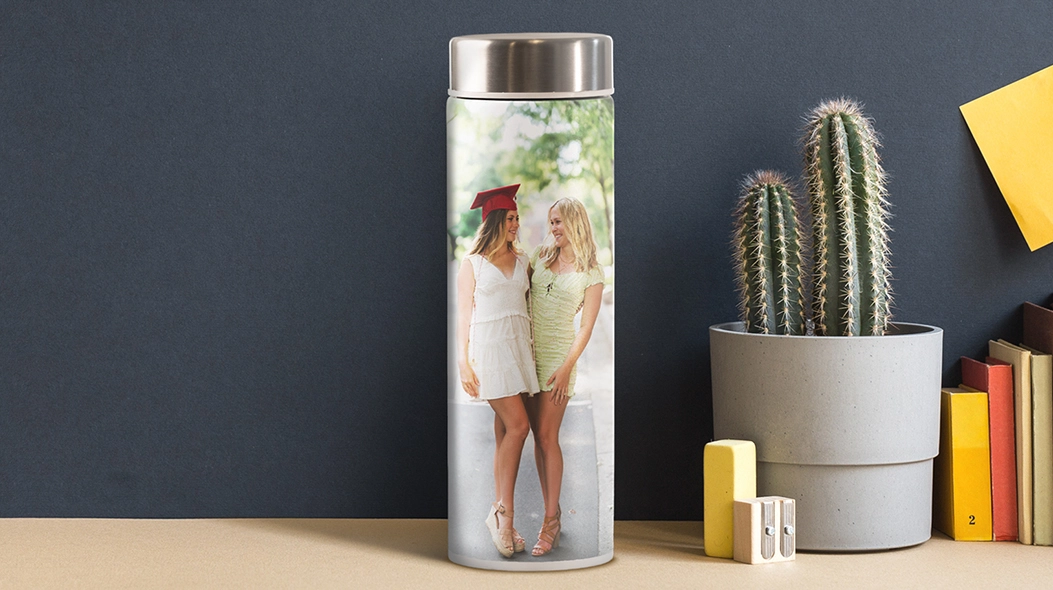 Educated And Hydrated: A matte white water bottle featuring a custom photo of two graduates, sitting on a desk next to a potted cactus and books.