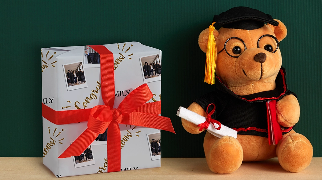 Wrap The Achievement: A gift box wrapped in custom graduation wrapping paper featuring photo collages and gold Congrats text, tied with a red ribbon next to a teddy bear wearing a graduation cap and gown holding a diploma.