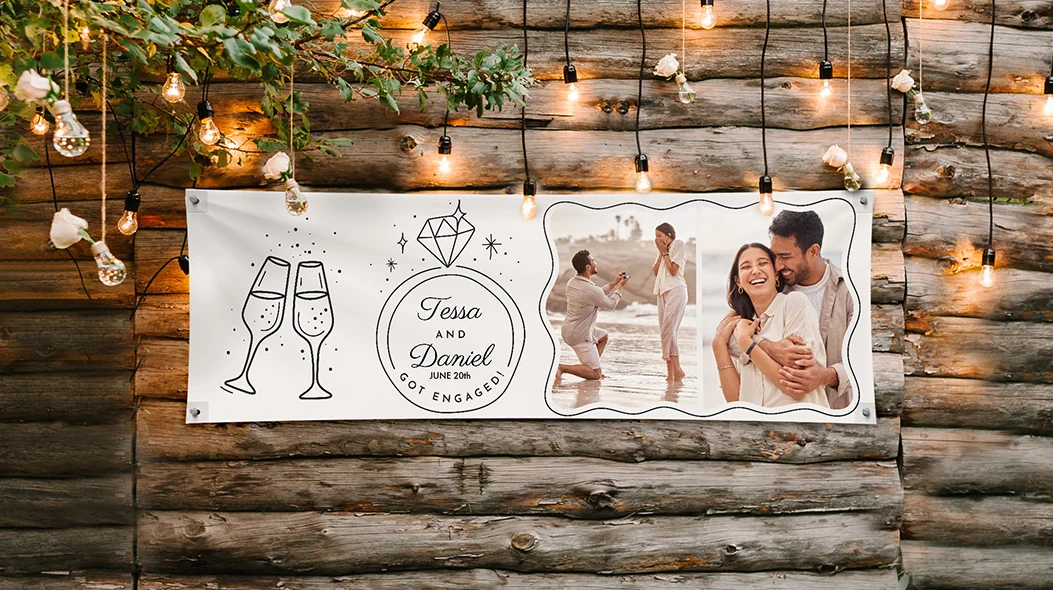 A white personalized horizontal banner for an engagement celebration hanging on a rustic wooden wall decorated with string lights, featuring illustrations of champagne flutes and a diamond ring alongside photos of a couple and the text Tessa and Daniel June 20th Got Engaged!